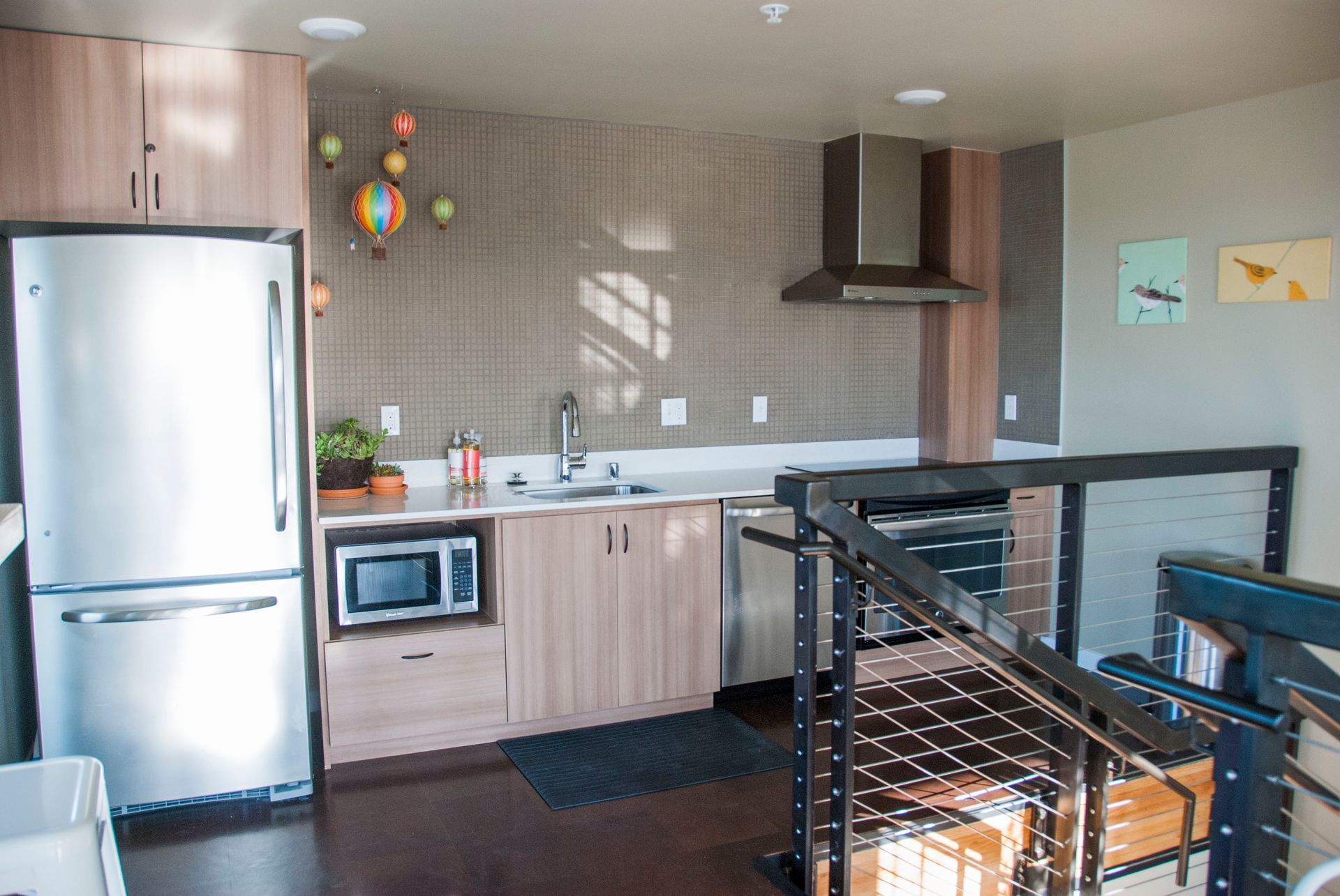 Modern kitchen with stainless steel fridge, light wood cabinets, white counters, and a staircase railing in foreground