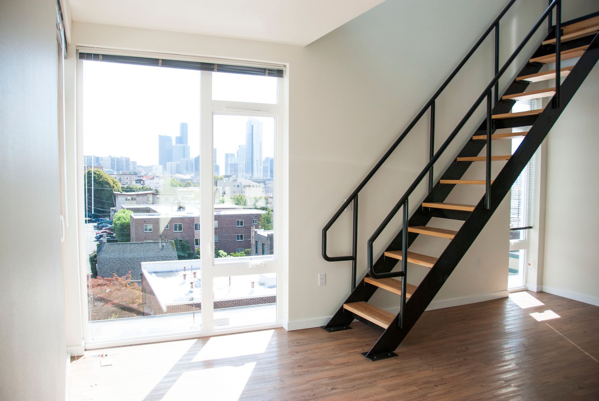 Bright apartment interior with black staircase, wood floors, and a balcony view of the city.