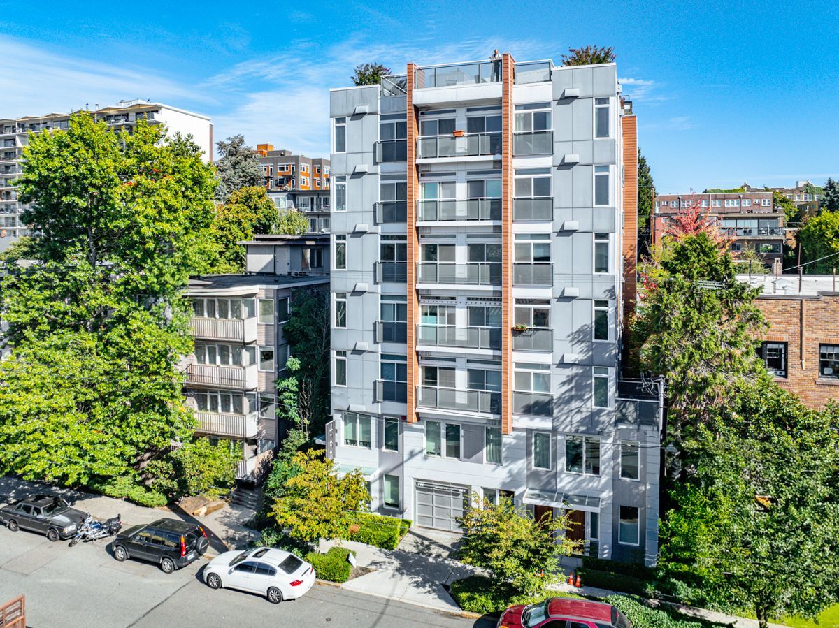 Modern apartment building with balconies, surrounded by trees and parked cars on a sunny street