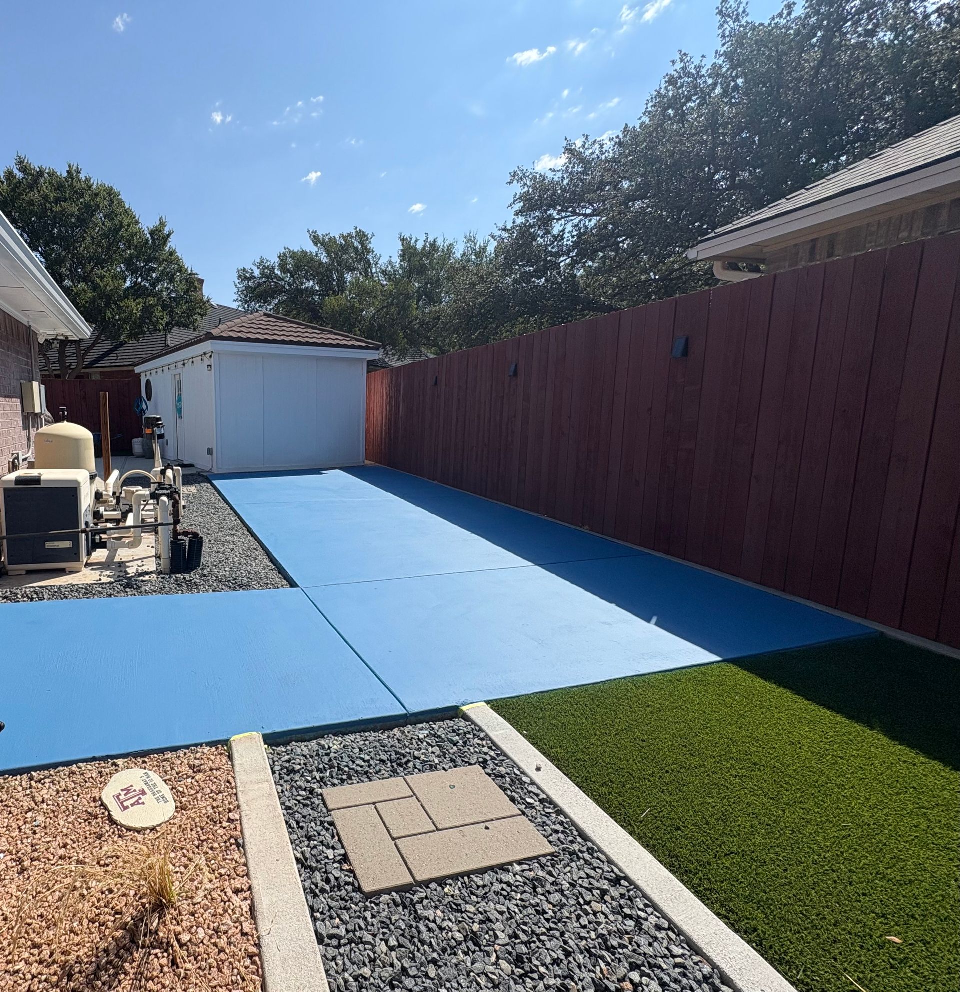 Blue mats in a backyard, adjacent to turf and a gravel path. Buildings and a red fence in the background.