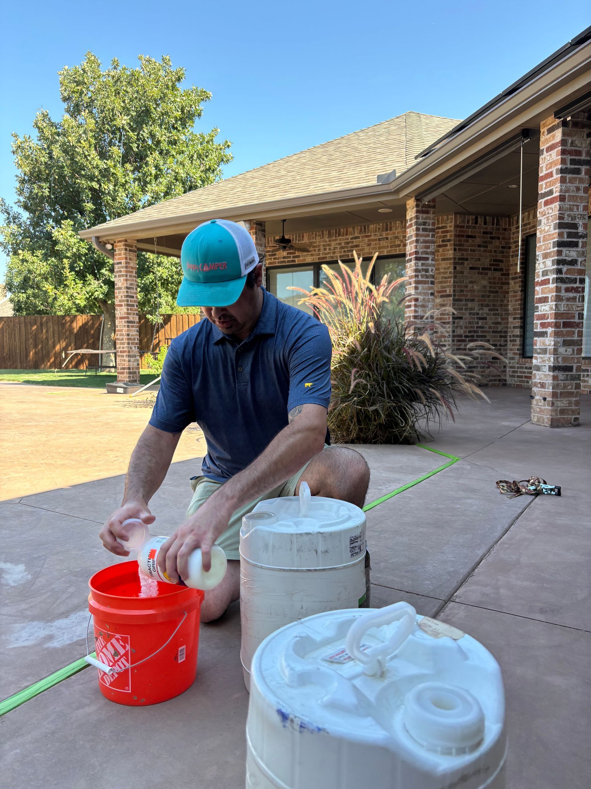 Man pouring liquid from a bottle into a red bucket, outdoors. He wears a hat and polo shirt.