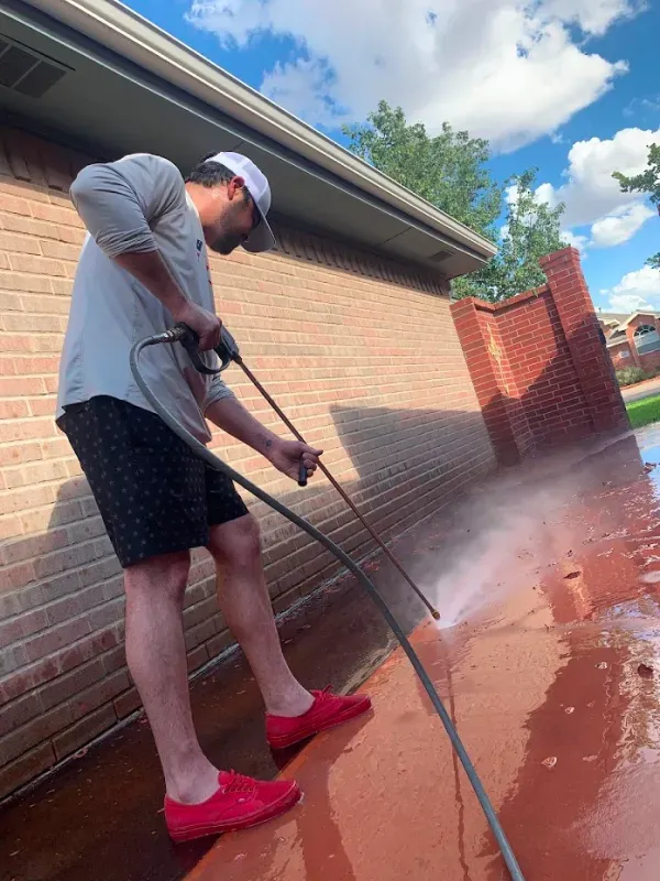 Man pressure washing a red-stained concrete patio near a brick wall, wearing red shoes, and a white cap.