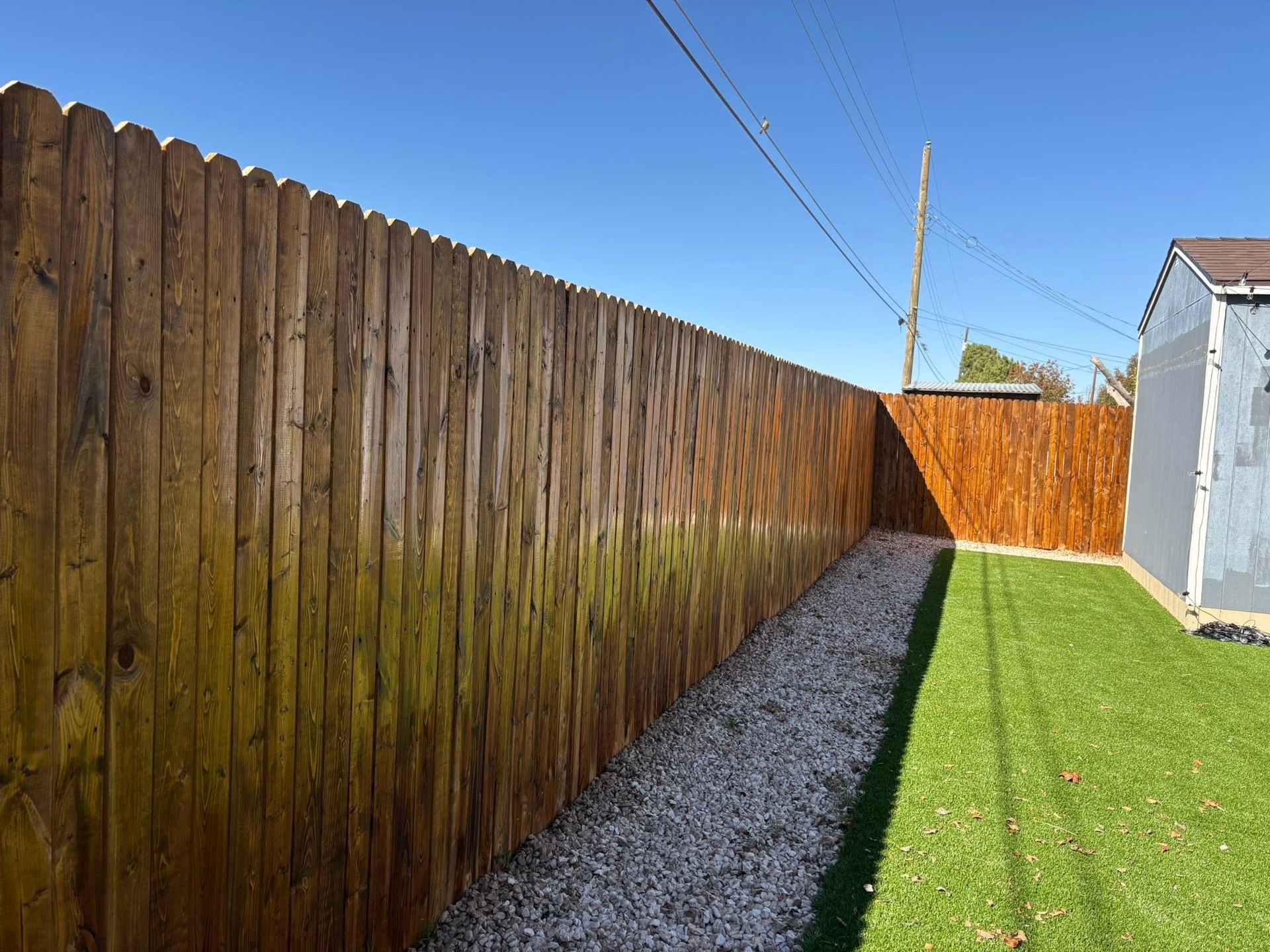 Brown wooden fence with a gravel path and green lawn in a sunny outdoor setting.