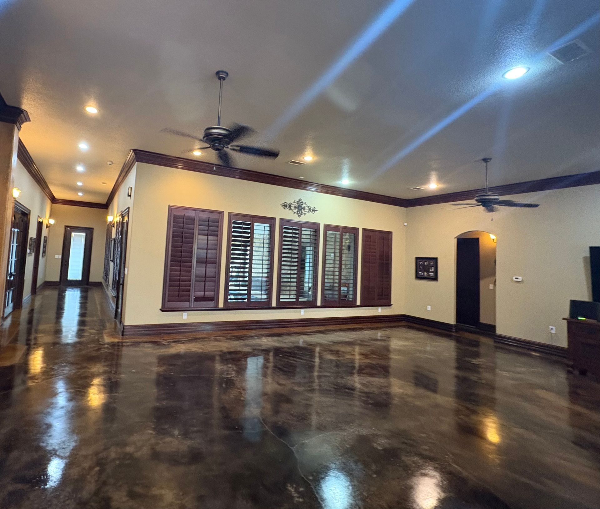 Spacious room with dark glossy floor, ceiling fans, and shuttered windows. Warm lighting and dark wood trim.