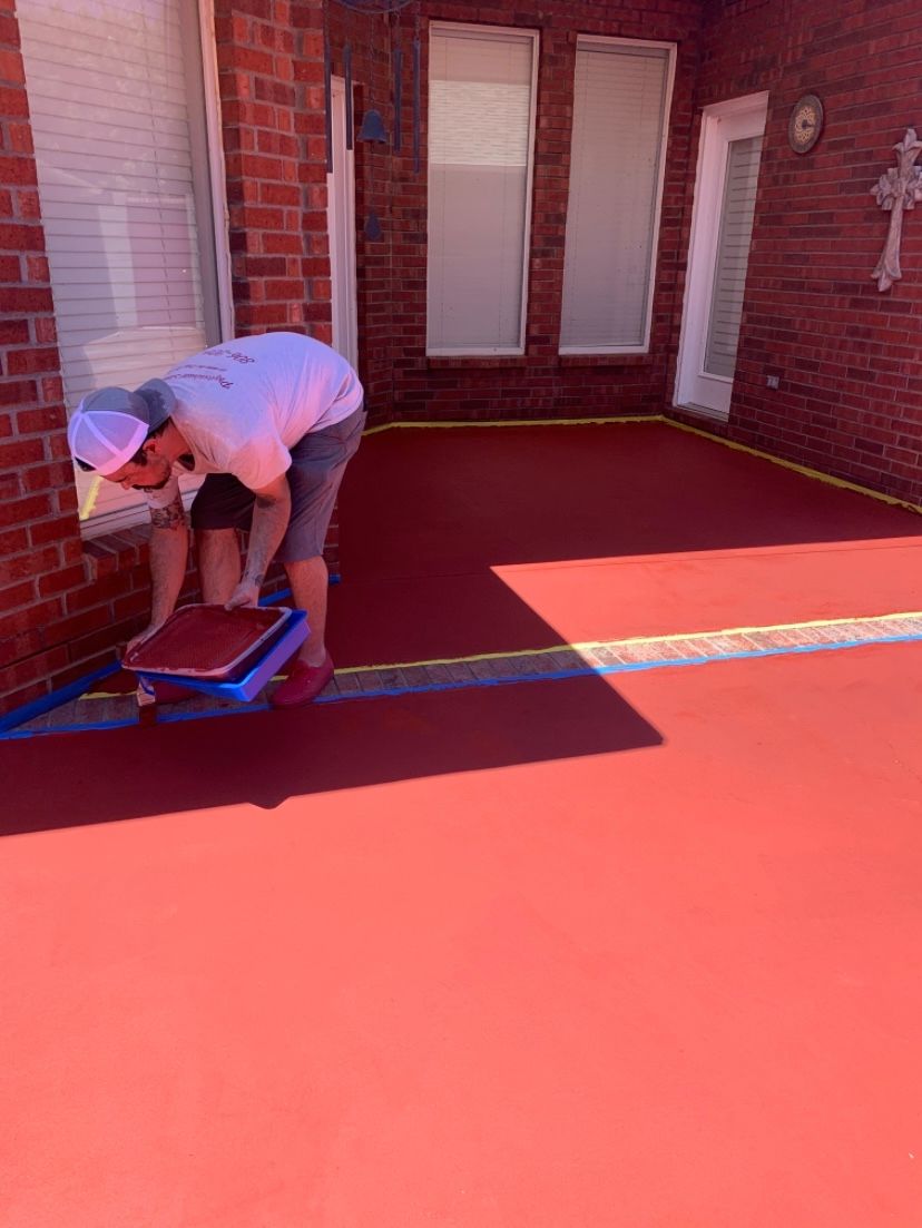 Man painting red concrete patio with a roller; brick building exterior.