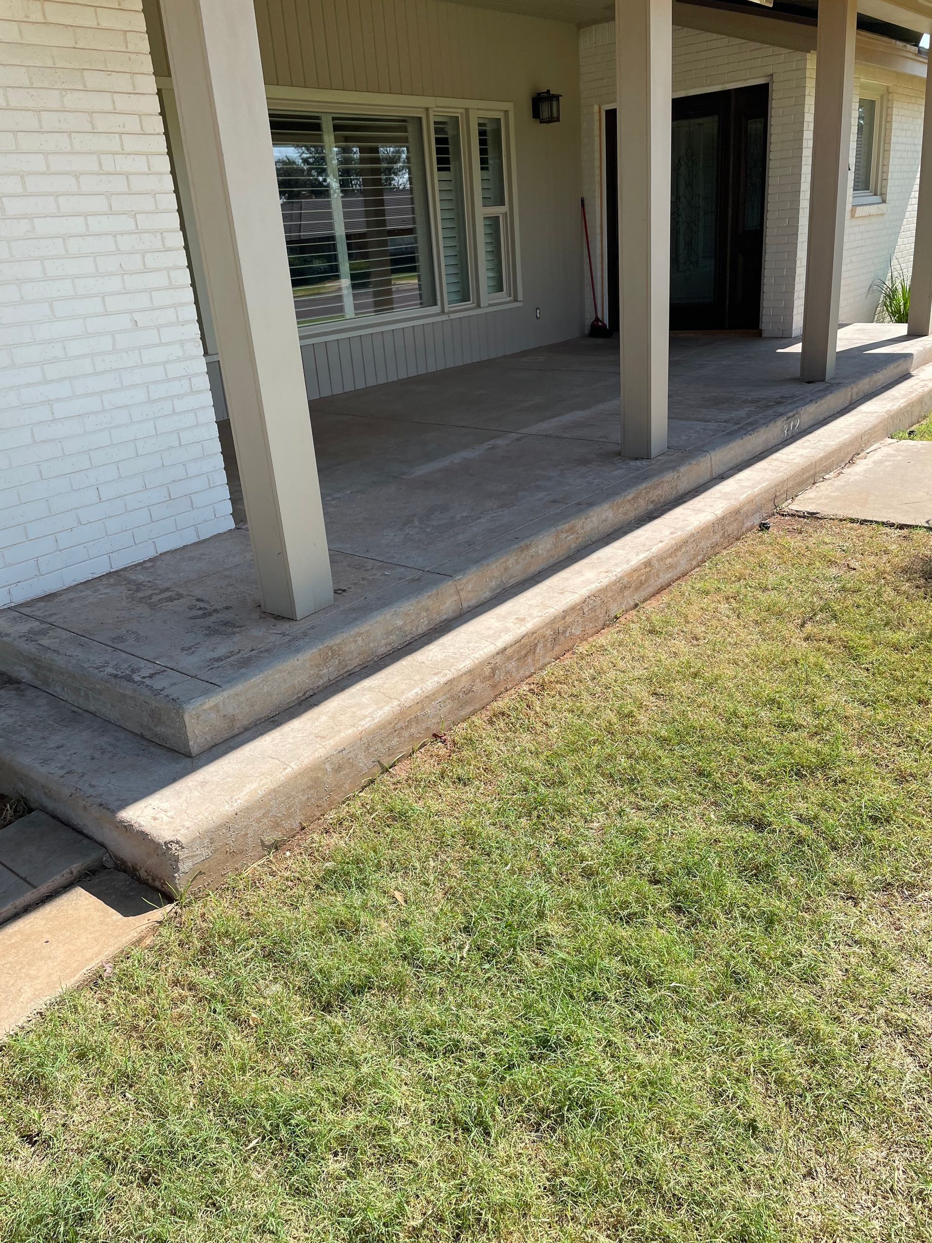 Front porch with concrete steps and support columns next to a brick wall and grassy lawn.