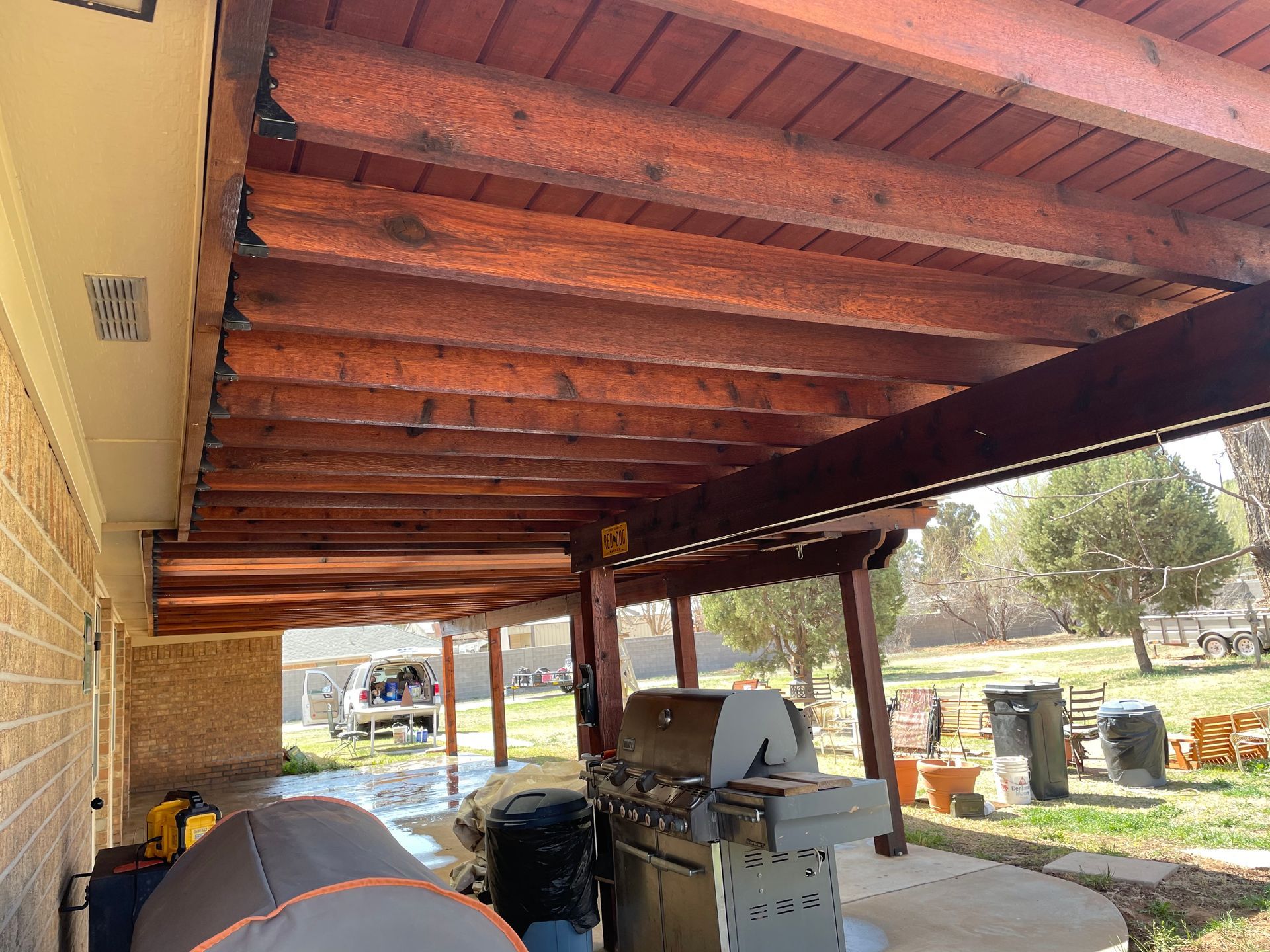 Wooden pergola attached to a brick home, with a grill and patio furniture beneath it.