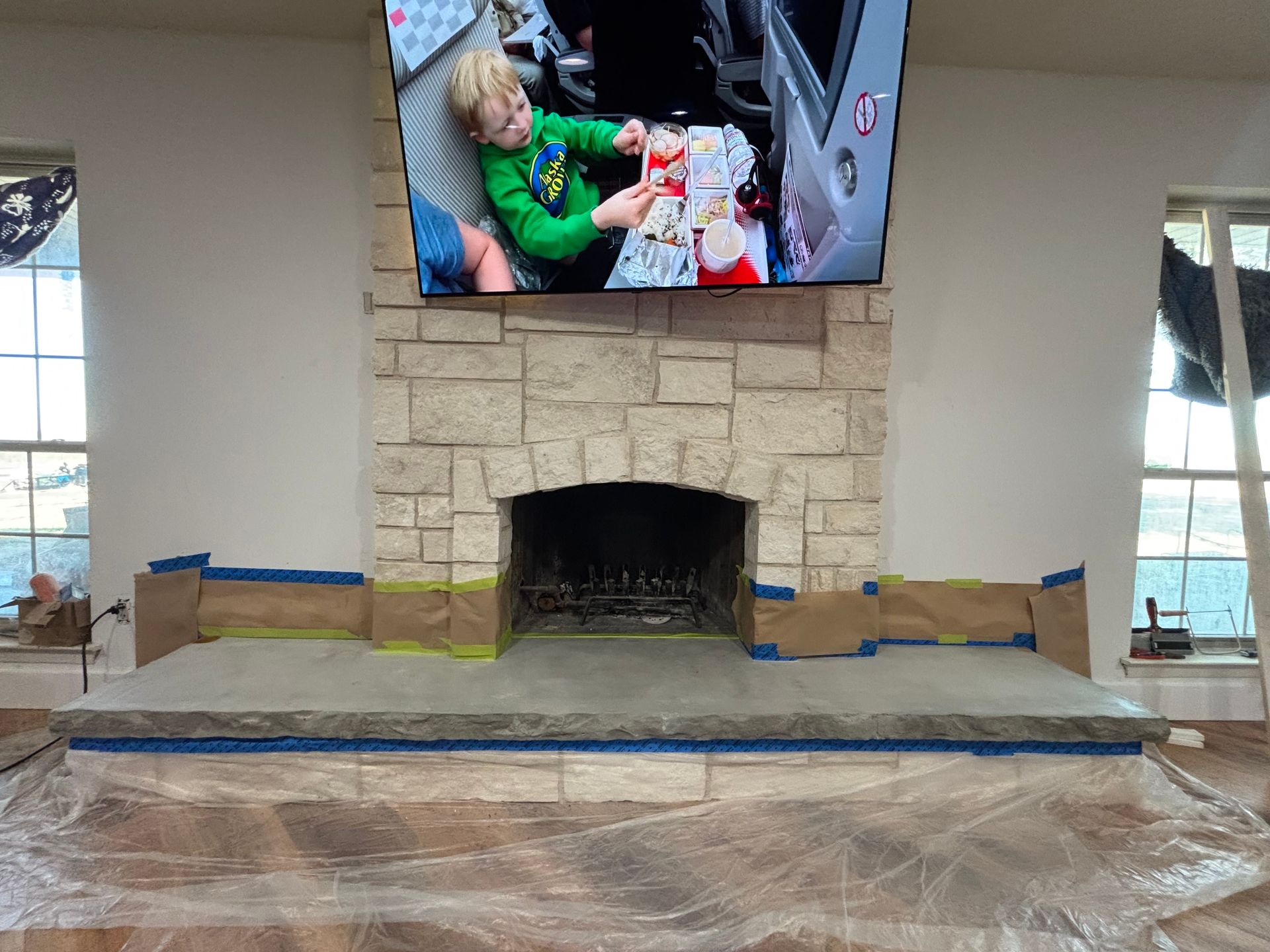 Fireplace with a stone facade, TV above, and a young child visible on the screen. The hearth is prepared for renovation.
