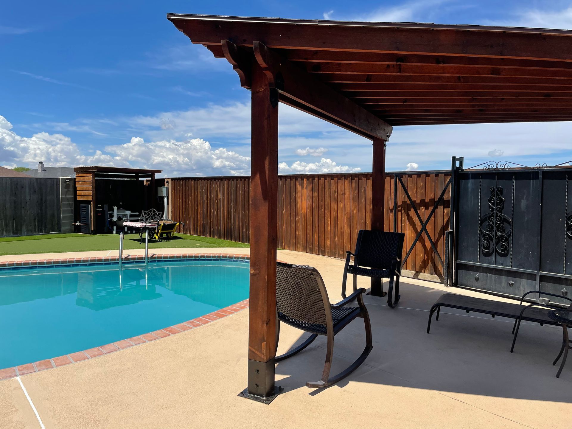 A backyard with a pool, pergola, wooden fence, and seating. Blue sky with clouds.