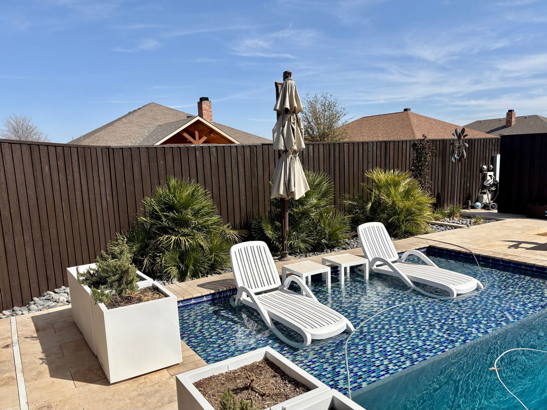 Swimming pool with lounge chairs, umbrella, plants, and brown fence under a blue sky.