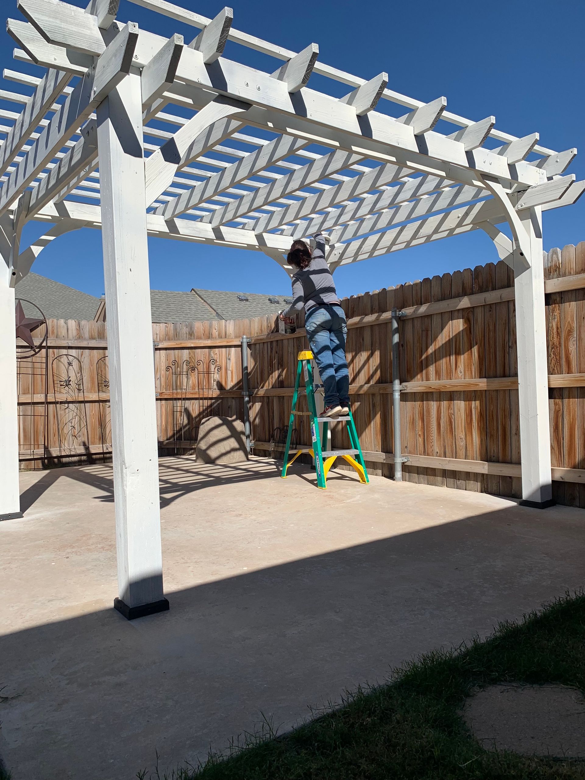 Person on a ladder, working on a white pergola. The pergola is in a concrete patio next to a wooden fence.