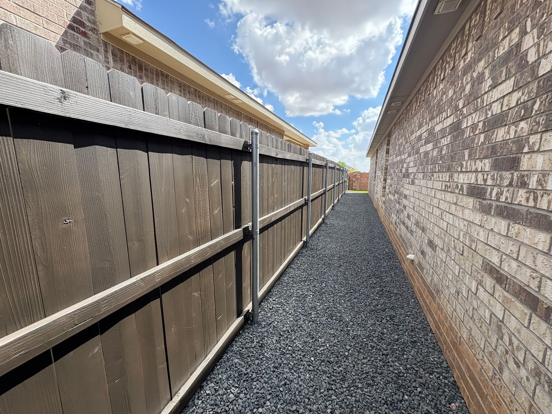 Narrow pathway between a brick building and a wooden fence, filled with dark gravel, under a blue sky.