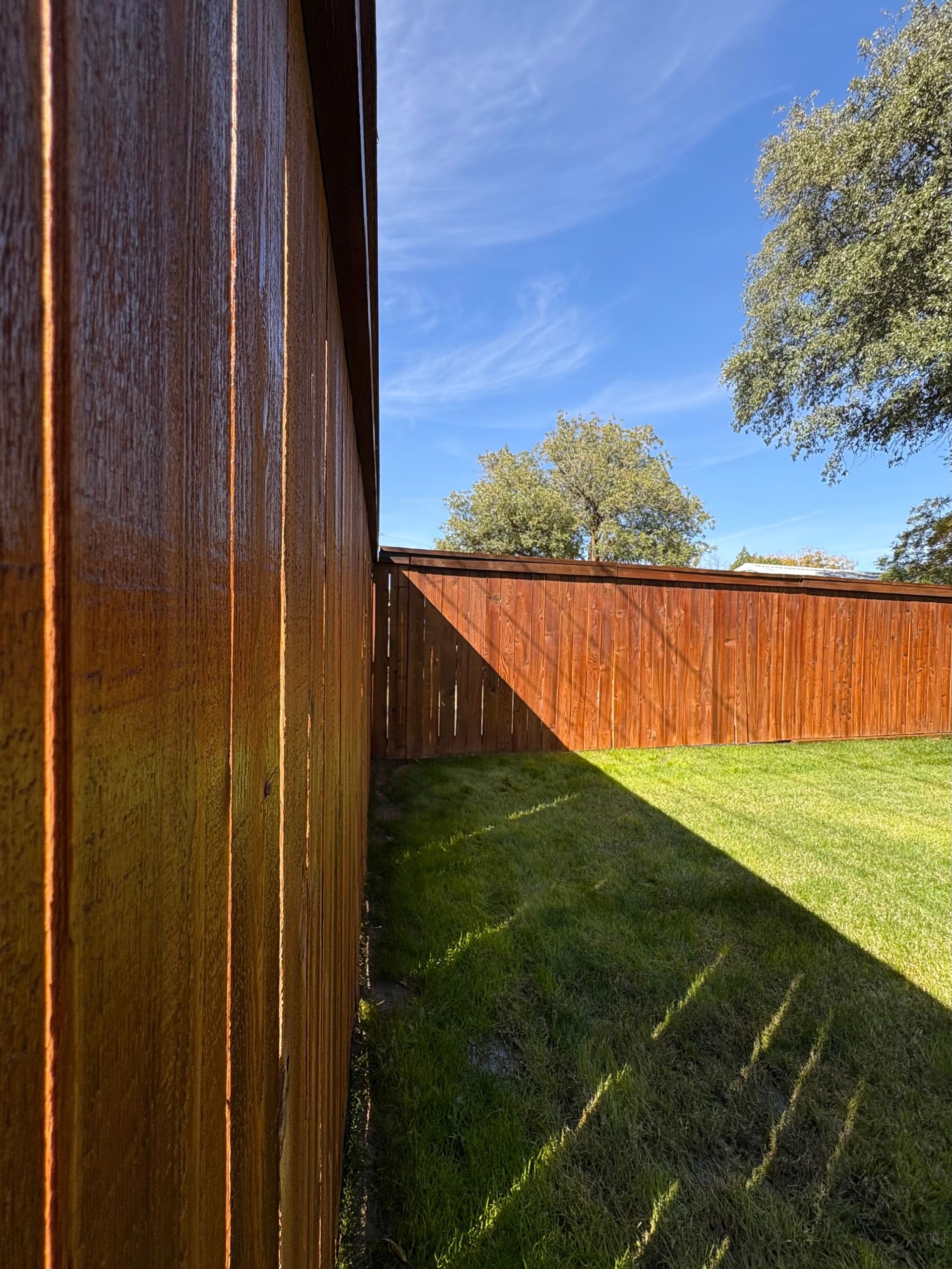 Wooden fence in a backyard with a grassy lawn, casting shadows under a blue sky.