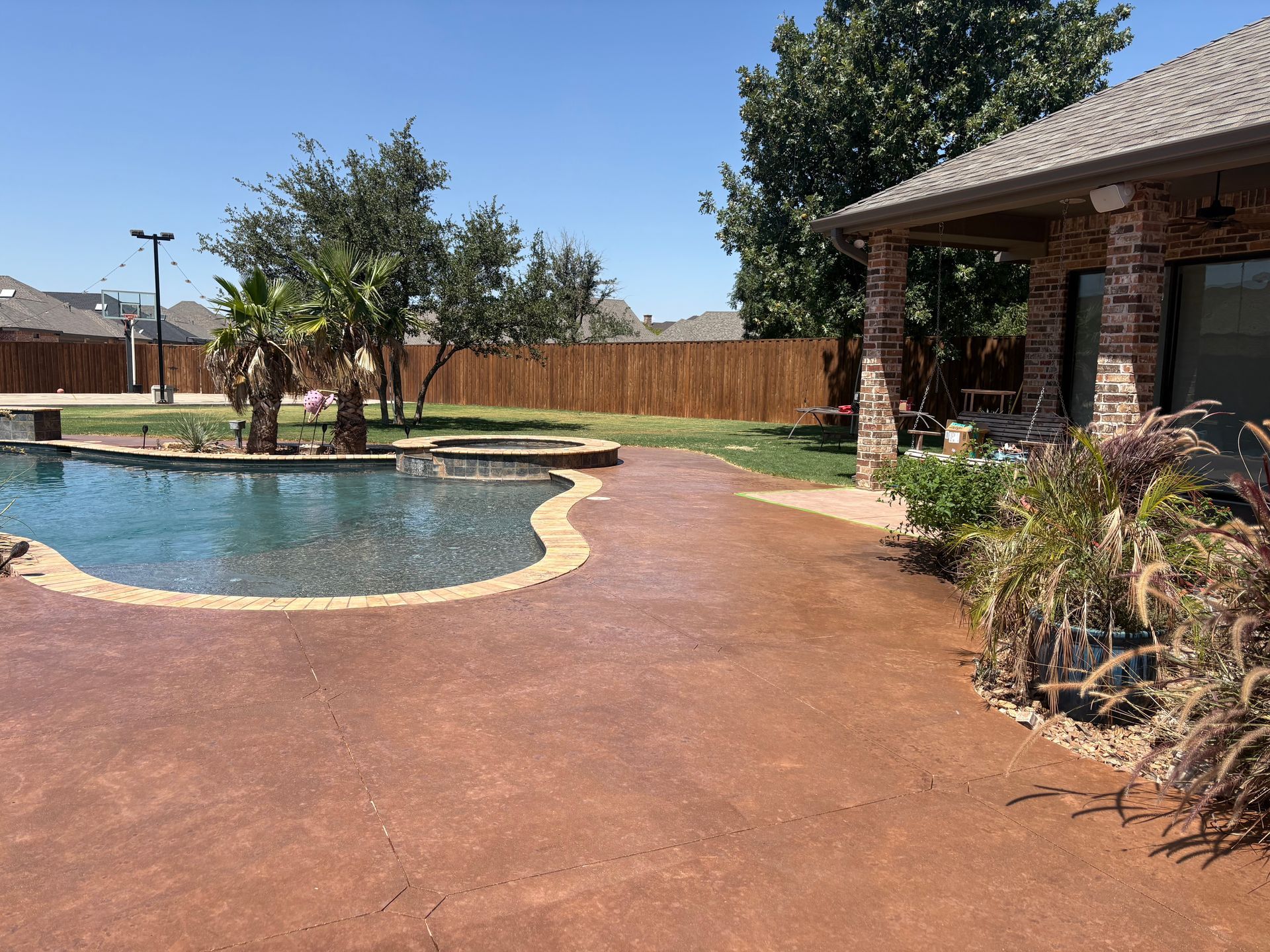 Backyard with pool and brick house; brown concrete patio, green lawn, trees, blue sky.