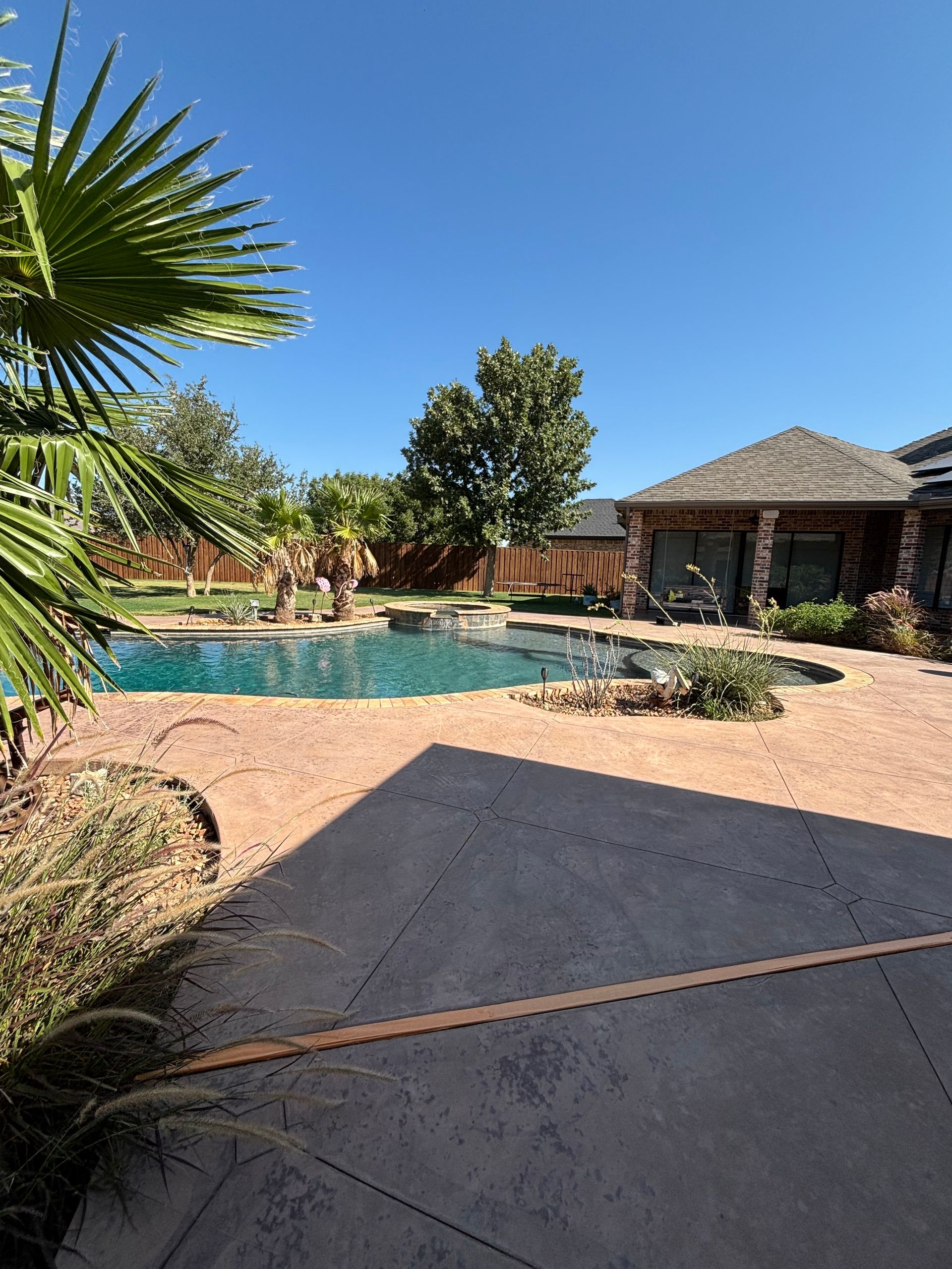 Backyard with a swimming pool, patio, and cabana under a blue sky. Palm trees and other greenery surround the pool.