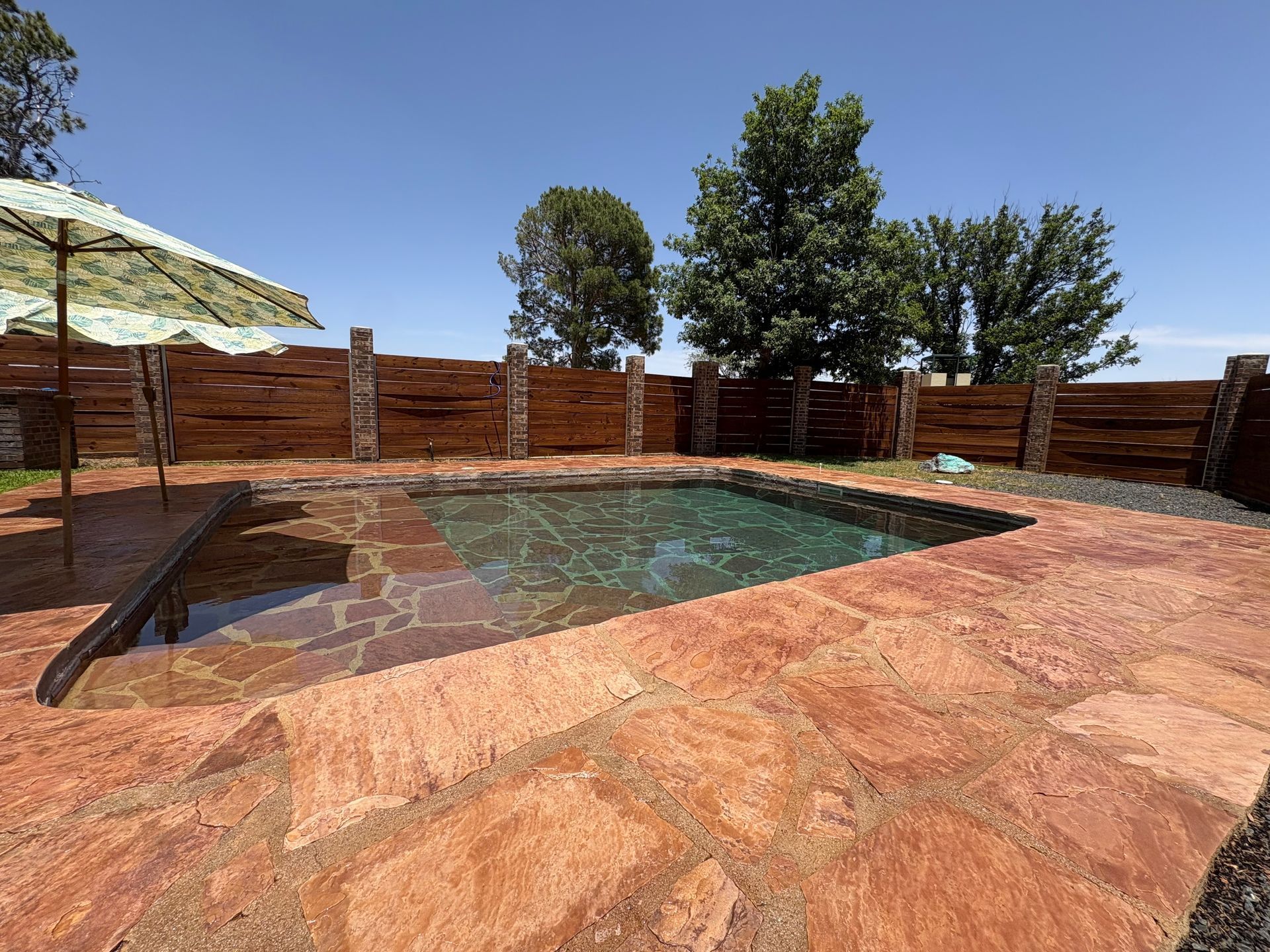 A small outdoor pool with brown stone surround, umbrella, and wooden fence on a sunny day.