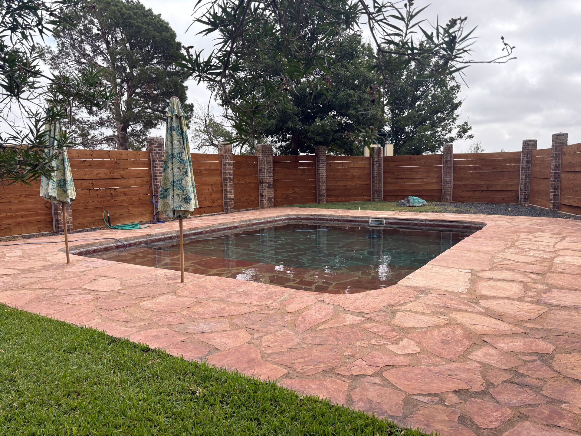Swimming pool surrounded by stone patio, lawn, and wooden fence; two umbrellas are open beside the pool.