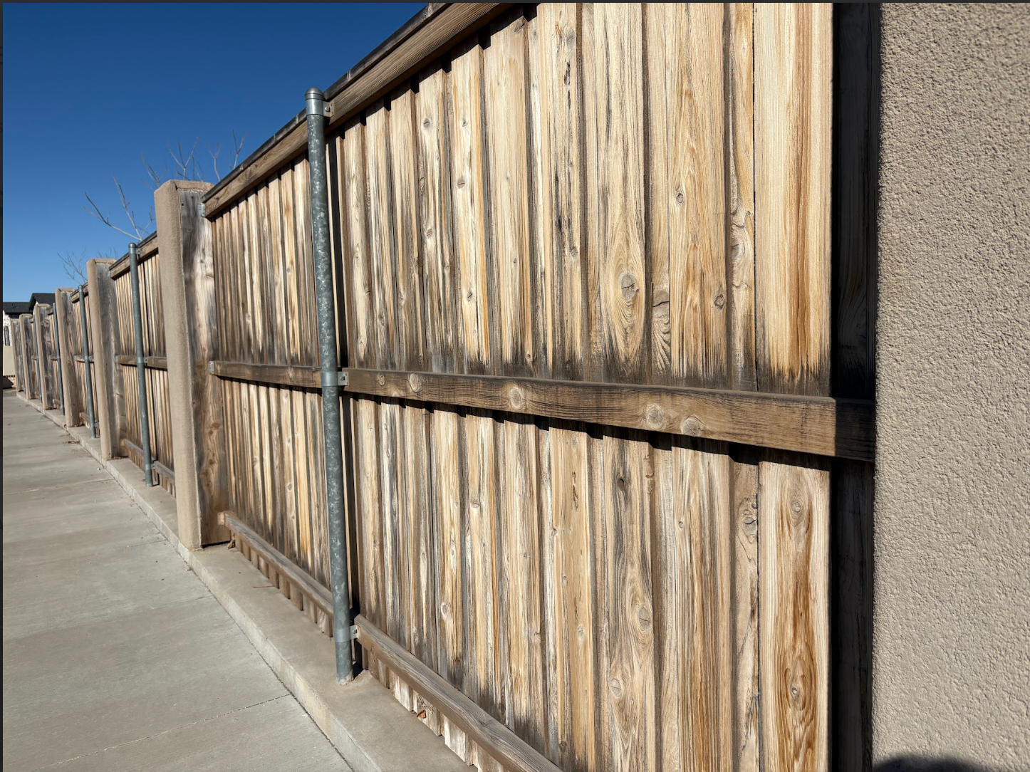 A weathered wooden fence with a metal support pole runs alongside a concrete sidewalk and a tan stucco wall.