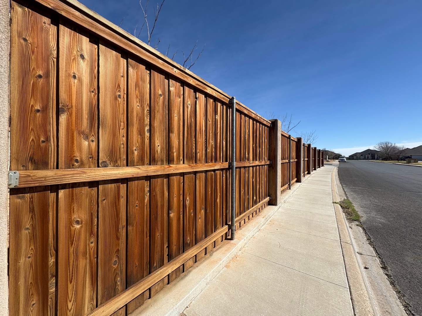 A tall, stained-wood fence runs alongside a concrete sidewalk and a paved road under a clear blue sky.