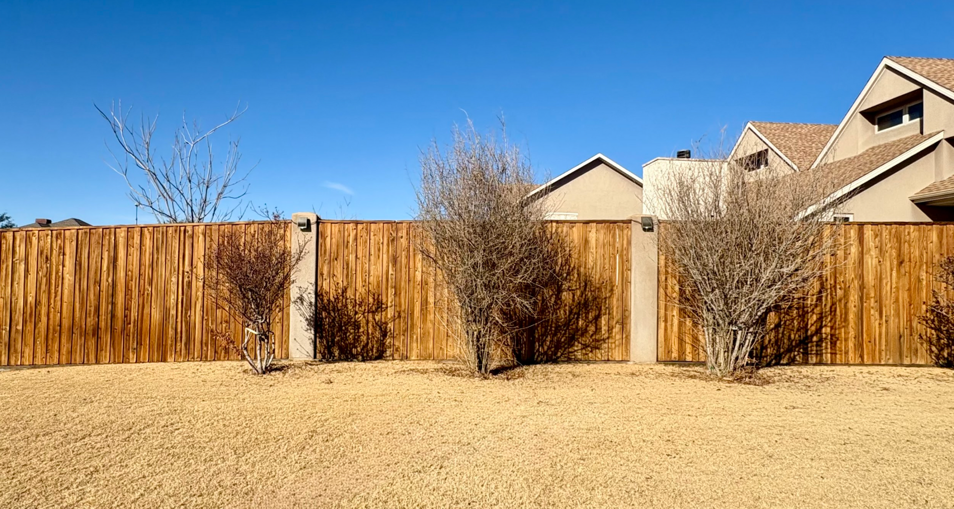 A tall wooden fence with concrete pillars in front of houses under a clear blue sky on a sunny day.