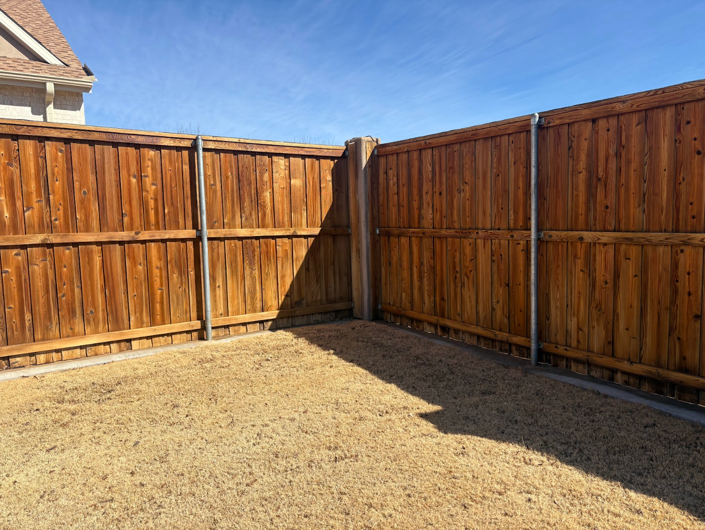 A corner of a backyard with a brown wooden privacy fence and a gravel ground area under a clear blue sky.