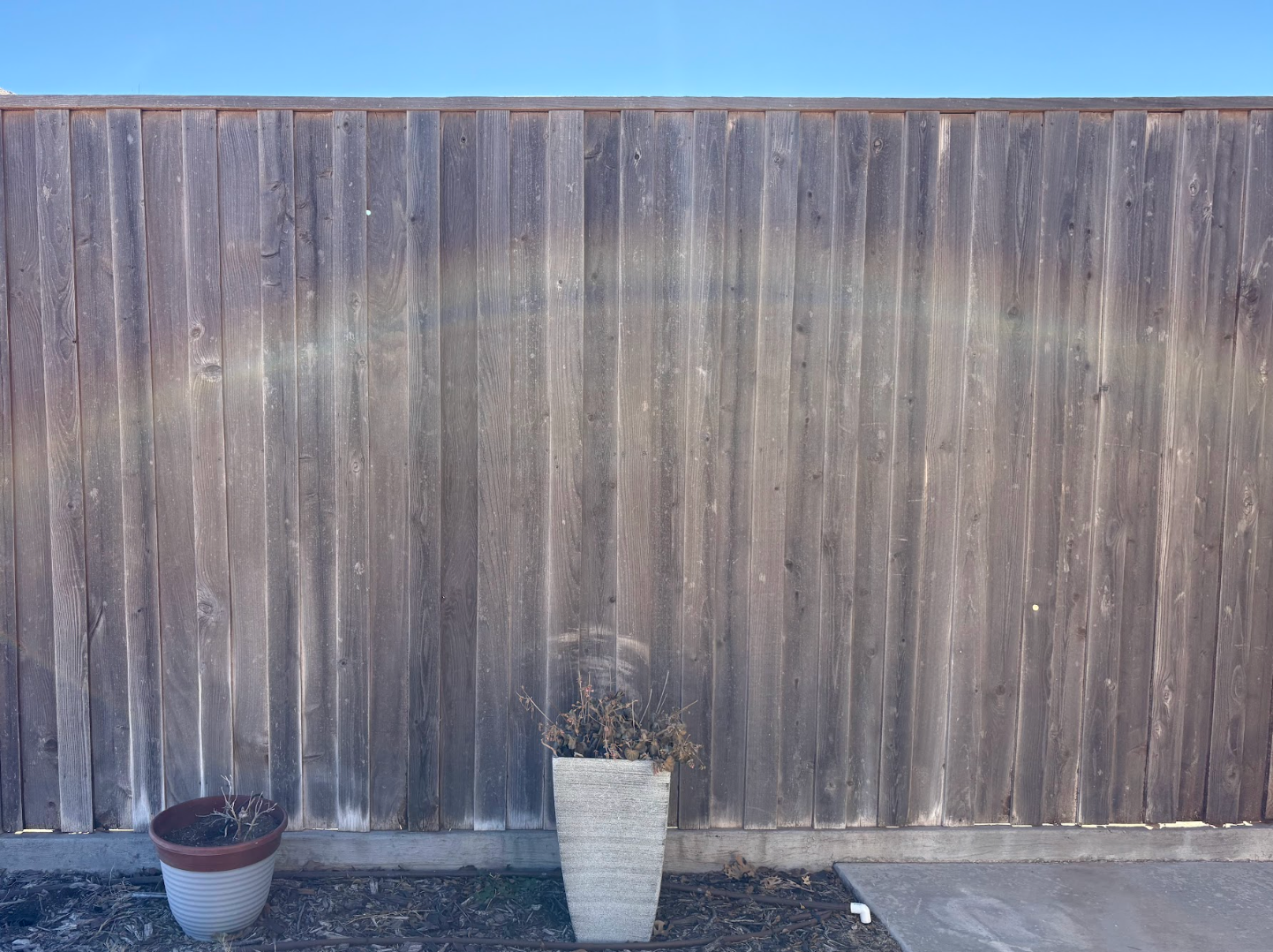 A faint, curved rainbow arc cast across a weathered wooden fence above two plant pots on a gravel ground.
