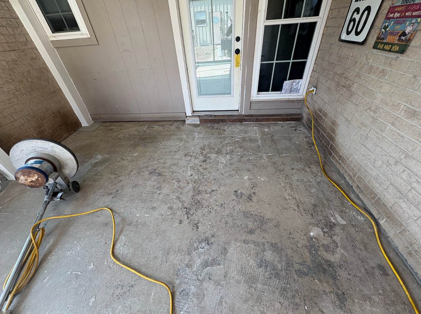 A floor buffer sits on a dirty concrete porch near a white door, with a yellow power cord snaking across the ground.