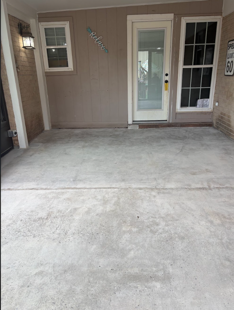 A concrete patio leading to a white front door, flanked by two windows on a brown wall with an outdoor light fixture.