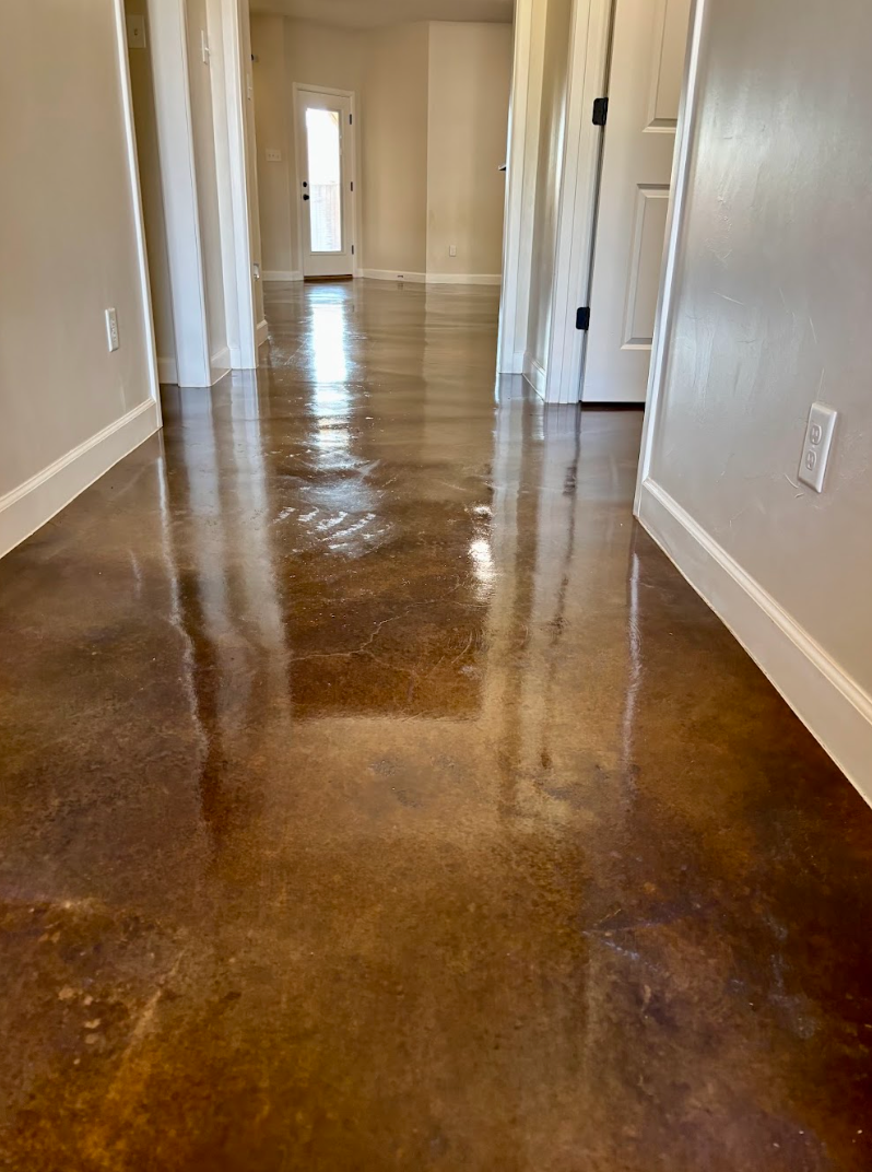 A polished, brown stained concrete floor in a bright, narrow hallway with white walls, baseboards, and a distant door.