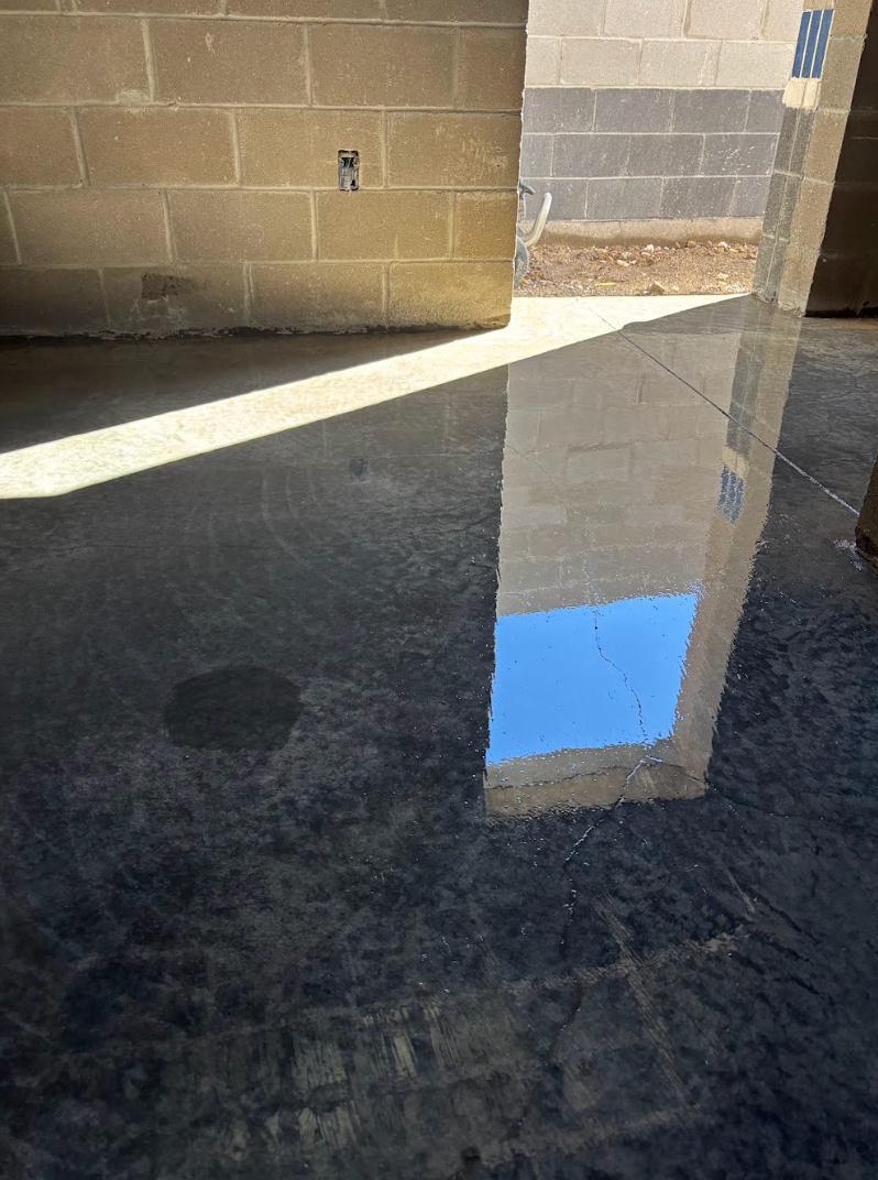 Polished dark concrete floor reflecting a bright blue sky and a block wall, bisected by a sharp diagonal beam of sunlight.
