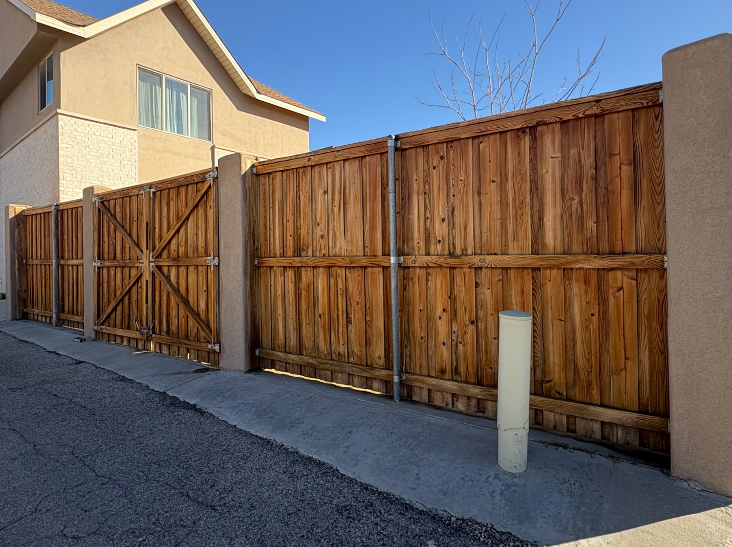 A wooden privacy fence with metal bracing stands between stucco pillars on a concrete path next to a suburban home.