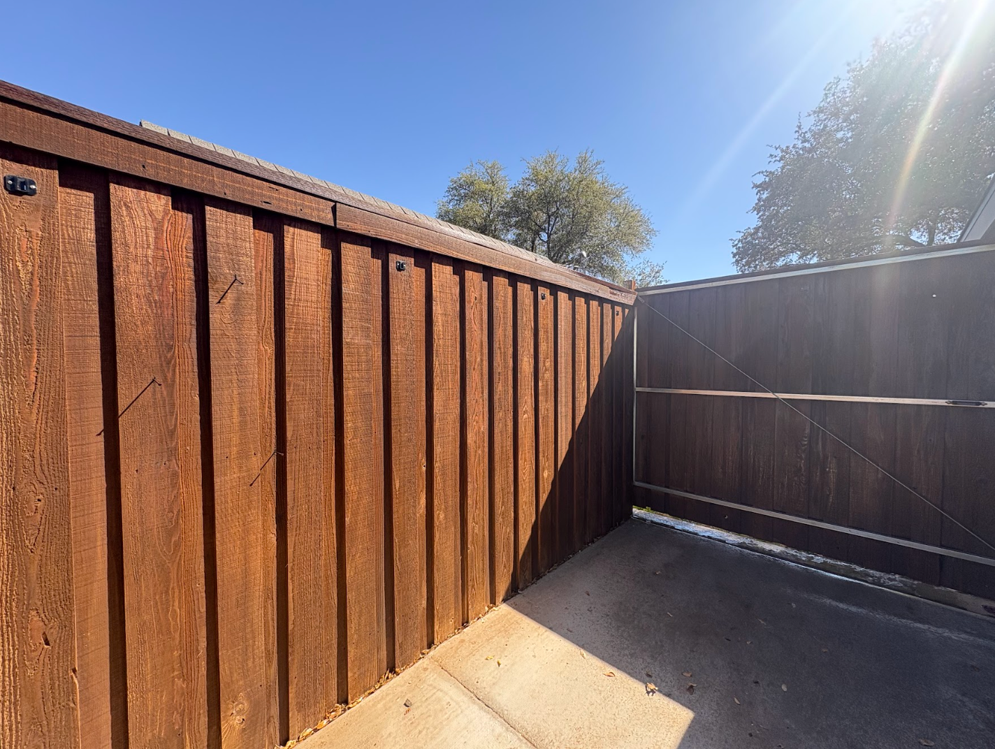 A corner view of a weathered brown wooden board-on-board fence meeting at a right angle above a concrete patio.