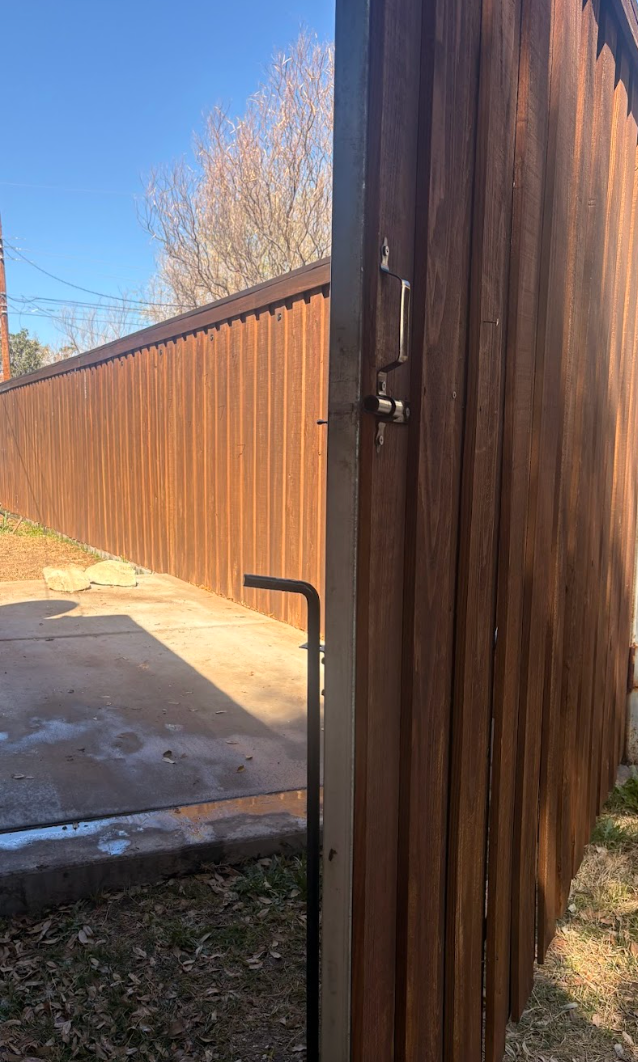A wooden privacy fence gate stands slightly ajar in a residential yard under a clear blue sky.