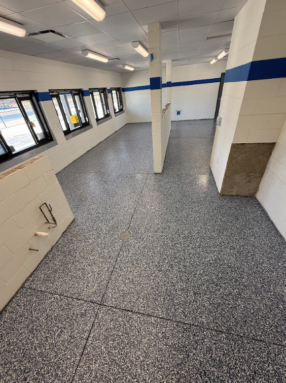 Empty indoor hallway with white walls, blue stripe, speckled floor, and windows on the left.