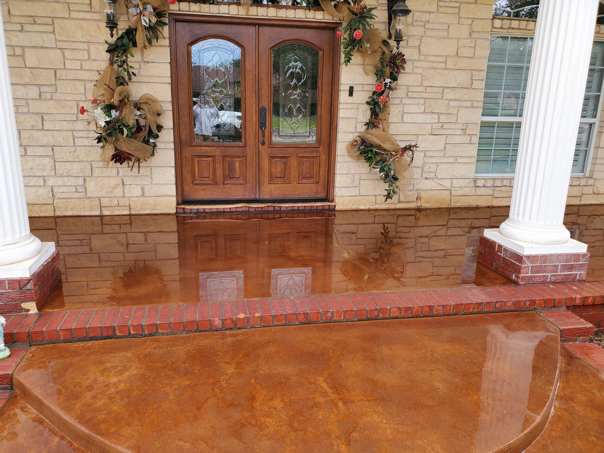 Covered patio with wet, brown concrete floor; red brick accents, wooden ceiling, and orange fence in background.