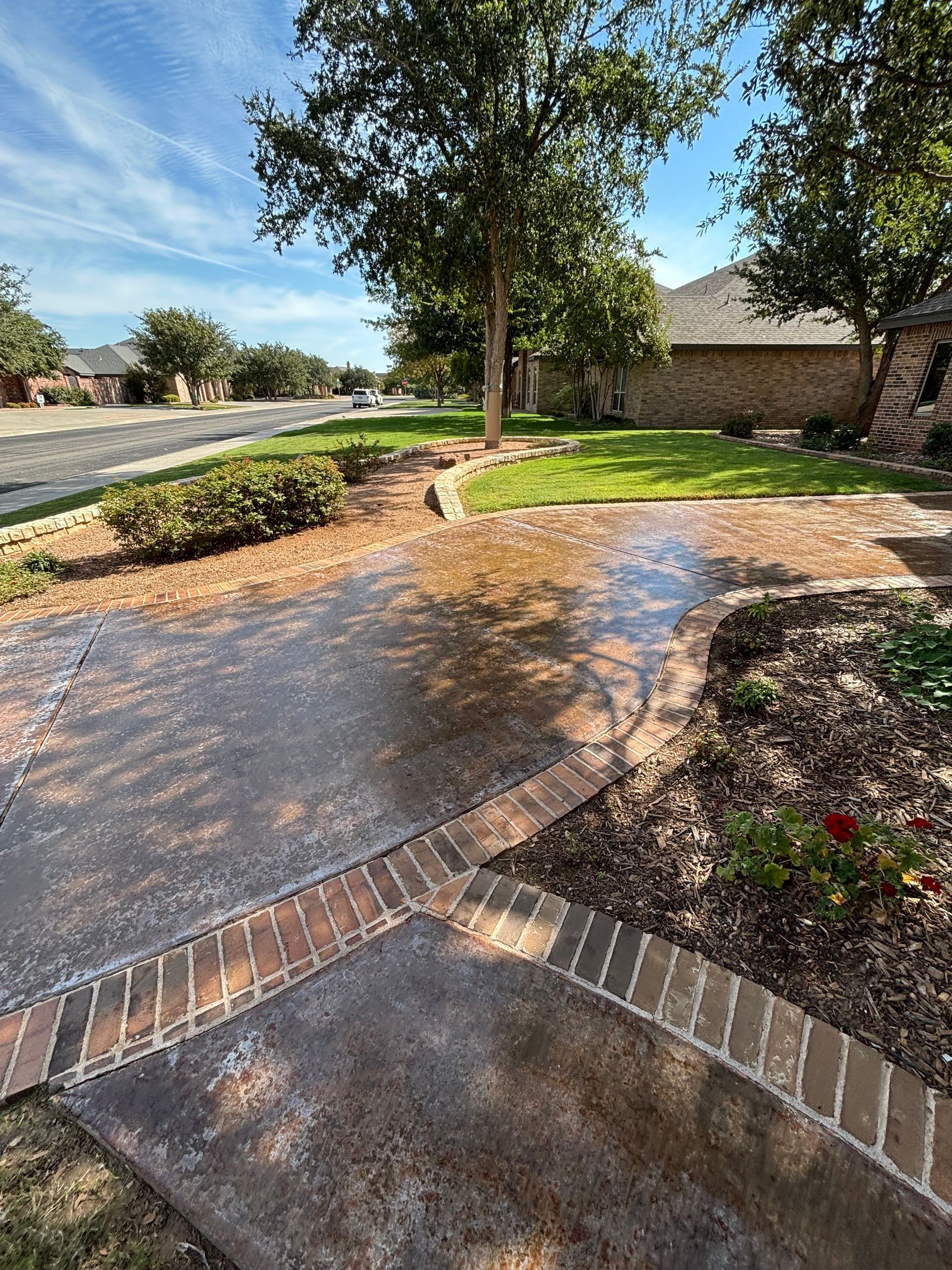 Driveway with brick border and landscaping, green lawn, tree, blue sky.