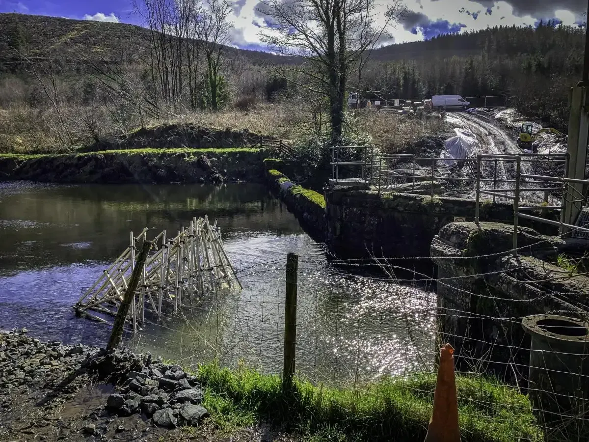 River with a wooden structure, possibly a dam, and a small bridge in a rural setting.