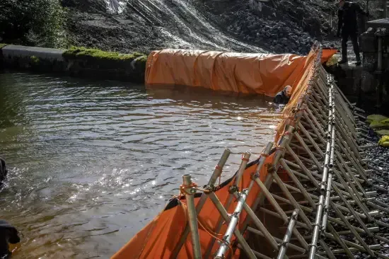 A temporary orange barrier made of fabric and bamboo is set up in a river, possibly for flood control or construction.