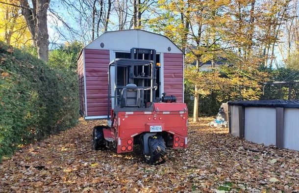 Un chariot élévateur rouge transporte un abri de jardin rouge et blanc dans une cour recouverte de feuilles, près d'arbres et d'une piscine.