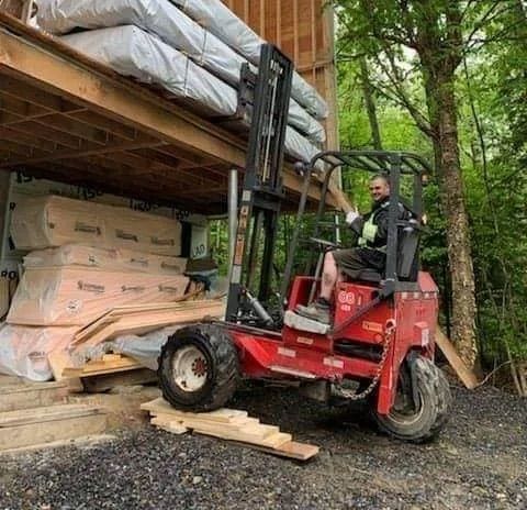 Un homme charge du bois sur un chariot élévateur sur un chantier de construction. Chariot élévateur rouge, bâtiment en bois et arbres.