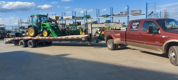 Une camionnette bordeaux tracte une remorque sur laquelle se trouve un tracteur vert. D'autres engins agricoles sont visibles.