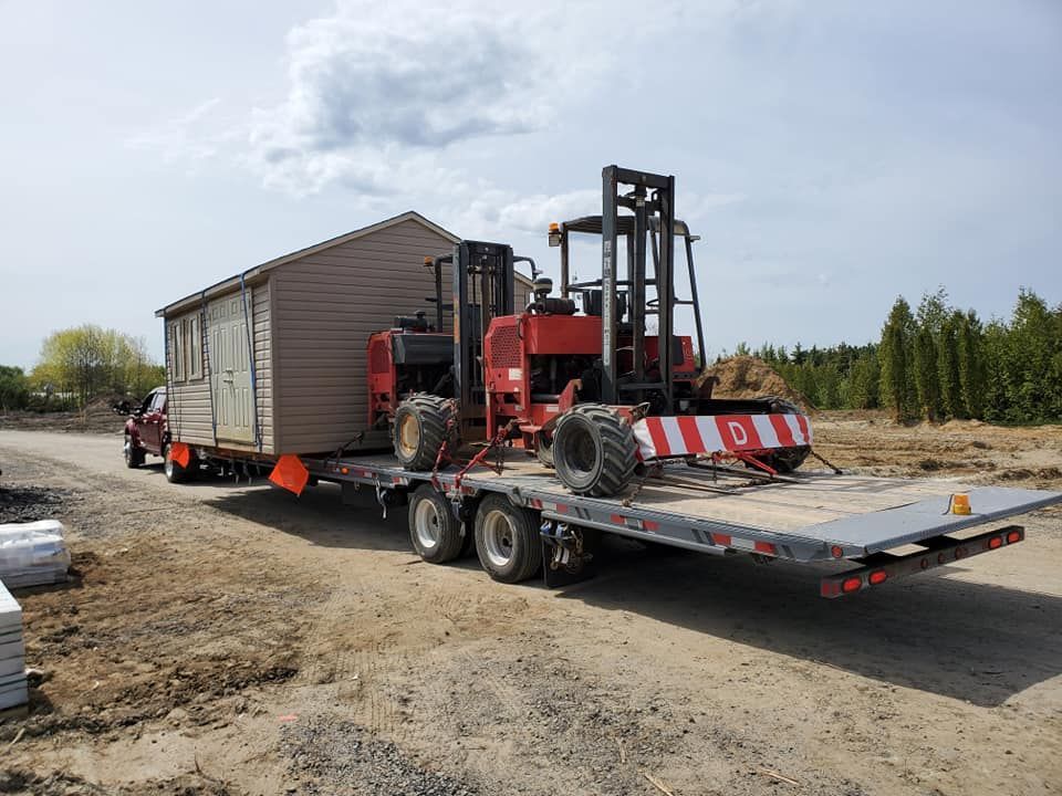 Un abri de jardin est transporté sur une remorque à plateau par un chariot élévateur rouge. Chemin de terre, journée ensoleillée.