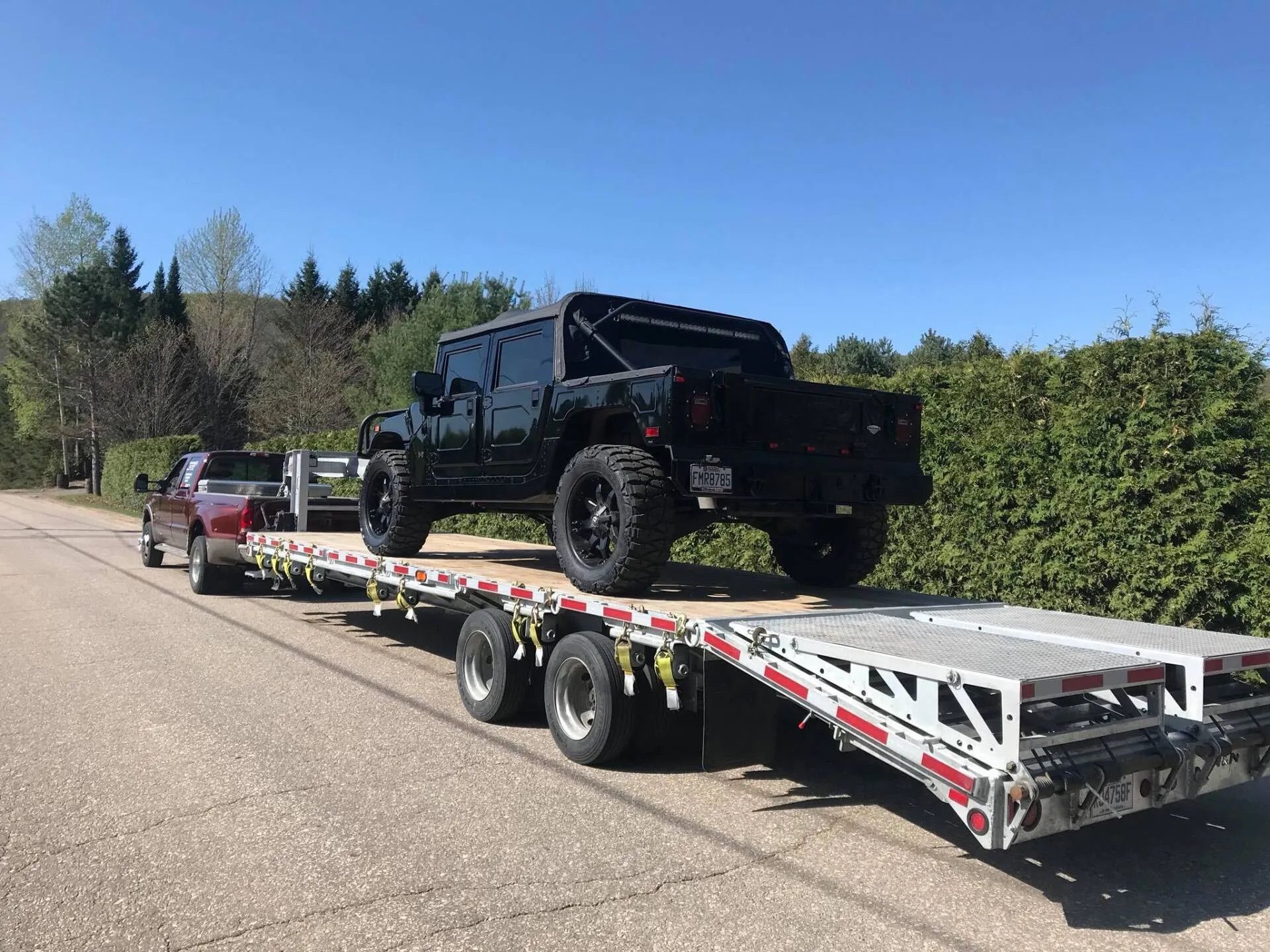 Black Hummer truck loaded on a flatbed trailer being towed by a red pickup truck on a paved road.