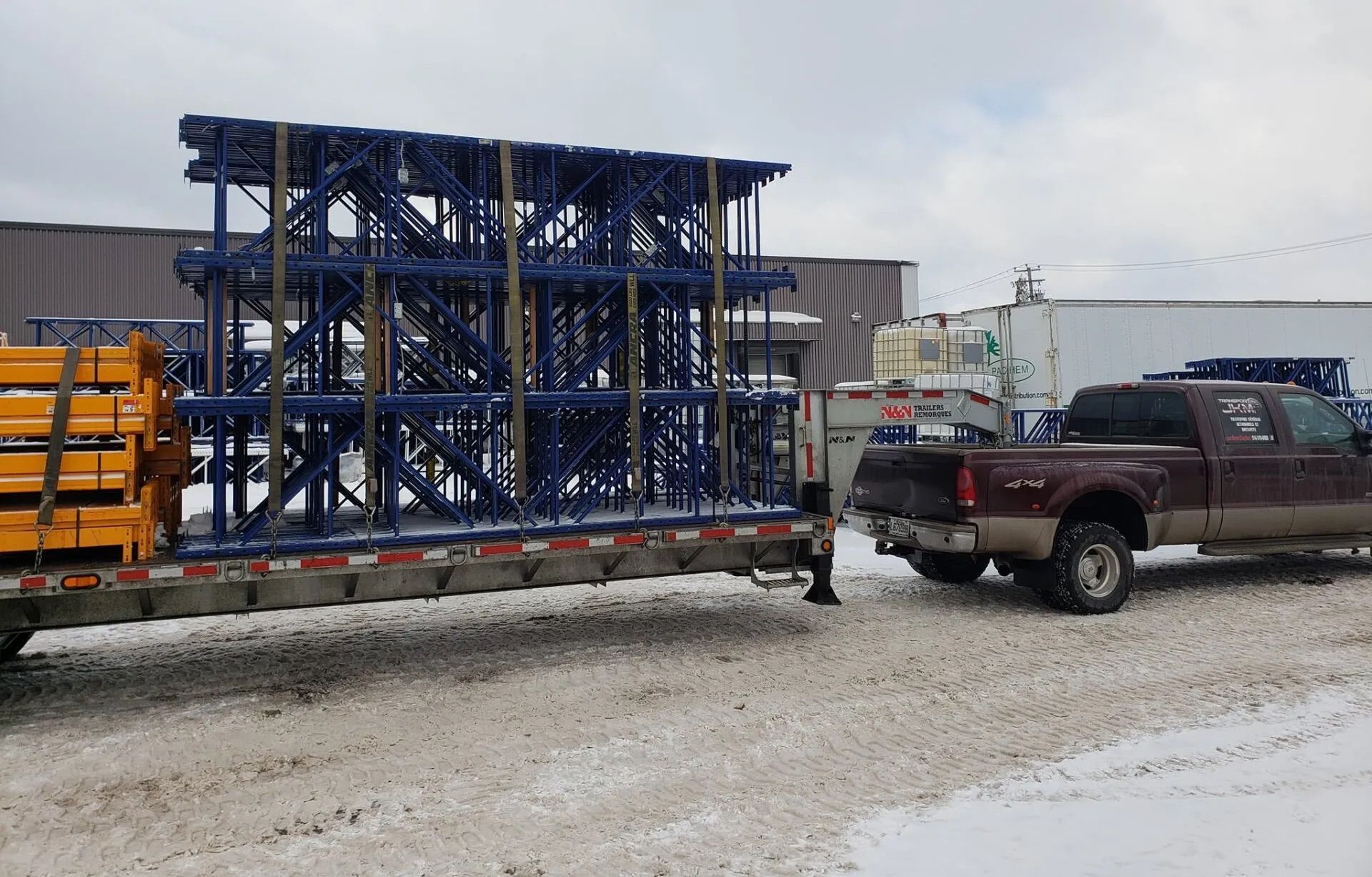 Des étagères métalliques bleues empilées sur une remorque à plateau, tirées par une camionnette marron par une journée enneigée.