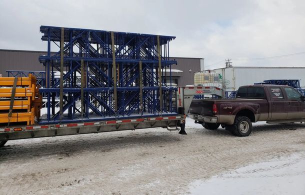 Des étagères industrielles bleues sont fixées sur une remorque à plateau, tirée par une camionnette marron par une journée enneigée.