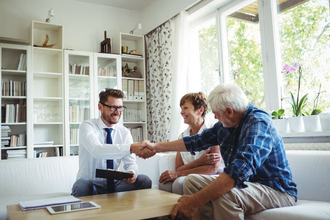 Man handing a document to two seated people in a bright living room with shelves and large windows