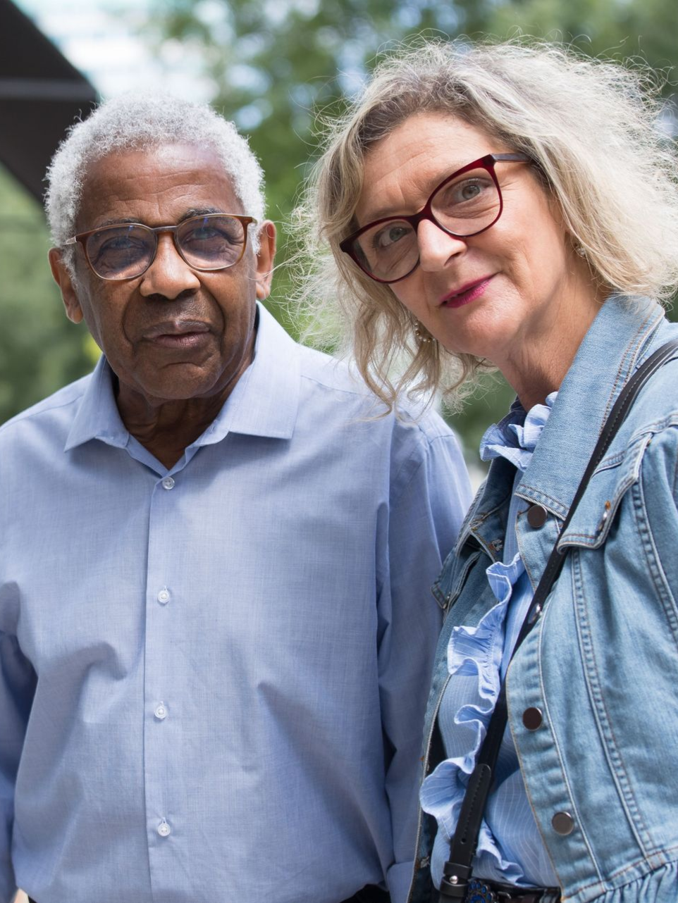 Two people standing outdoors, one in a blue shirt and one in a denim jacket, both facing the camera.
