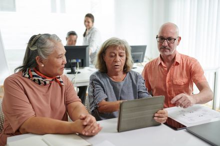 Three people review a tablet at a bright office desk with papers and a computer in the background.