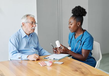 Doctor explaining medication to a patient at a clinic table, with pill packets and a prescription pad nearby