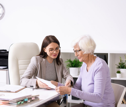 Two women reviewing documents together at an office desk, one pointing to a page, with a laptop and plants nearby.